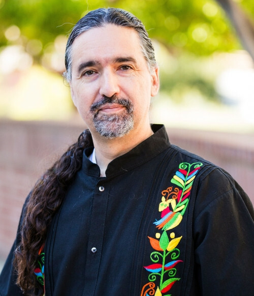 A photo of Dr. Knight: a middle-aged man with dark eyes and long dark hair worn in a ponytail, with a salt-and-pepper goatee. He is wearing a black guayabera shirt with two vertical colorful flower and bird accent stripes down from the shoulders. He is attempting to look friendly  and relatable, the sort of guy you could tell your problems too and get good observations and feedback from.